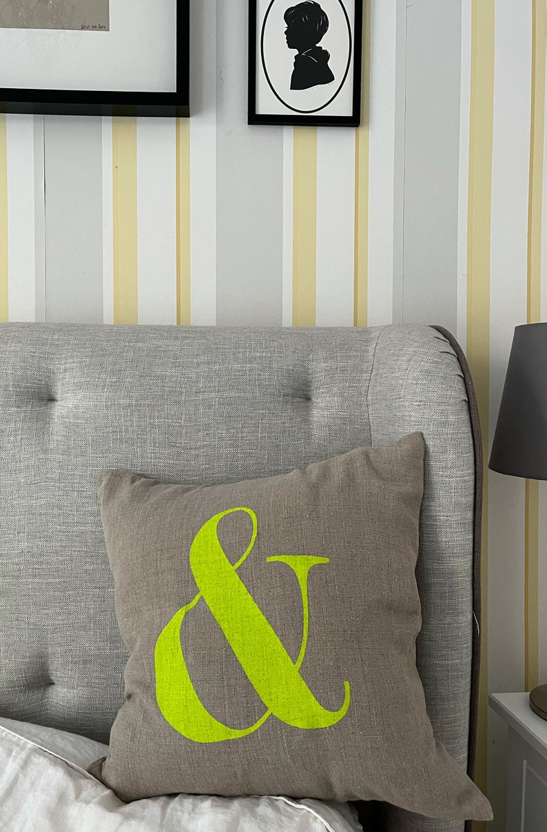 May include: A brown cushion with a neon green ampersand symbol, placed on a grey upholstered headboard. The background features striped wallpaper in white, yellow, and grey. The image also shows framed artwork.