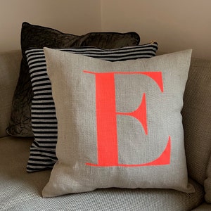 May include: A grey linen cushion with a large, bright pink letter "E" printed on the front. The cushion is sitting on a grey sofa.