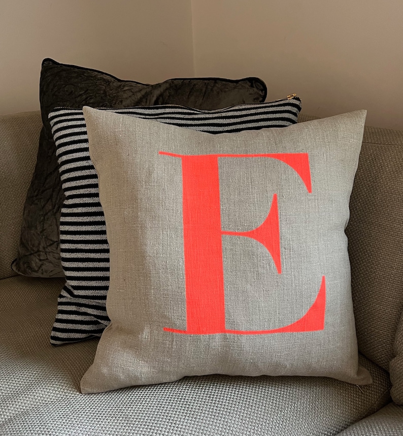 May include: A grey linen cushion with a large, bright pink letter "E" printed on the front. The cushion is sitting on a grey sofa.