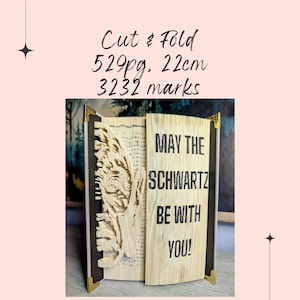 May include: An open book with pages cut and folded to reveal a detailed design. The text "MAY THE SCHWARTZ BE WITH YOU!" is visible on the right page. The book has gold corner protectors and measures 22cm in height.