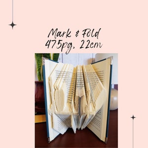 May include: A book with 475 pages has been folded into a 3D design. The book is 22 cm wide and has a dark blue cover. The book is sitting on a wooden table.