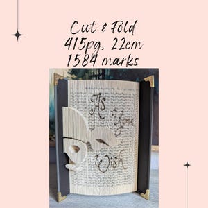May include: A decorative book with pages cut and folded to reveal a design and the words "As You Wish." The book has a black spine and gold corner protectors. The text "Cut & Fold, 415pg, 22cm, 1584 marks" is written at the top.