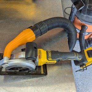 Puede incluir: Una sierra circular DeWalt amarilla y negra con una hoja plateada y un accesorio de recogida de polvo naranja. Una manguera negra conecta la sierra a una aspiradora de taller naranja y negra.
