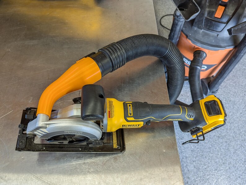 Puede incluir: Una sierra circular DeWalt amarilla y negra con una hoja plateada y un accesorio de recogida de polvo naranja. Una manguera negra conecta la sierra a una aspiradora de taller naranja y negra.