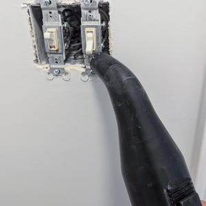 Puede incluir: Dos interruptores de luz instalados en una pared, con cableado expuesto. Una boquilla negra apunta hacia los interruptores, posiblemente para limpieza o reparaci&oacute;n. La pared est&aacute; pintada de blanco.