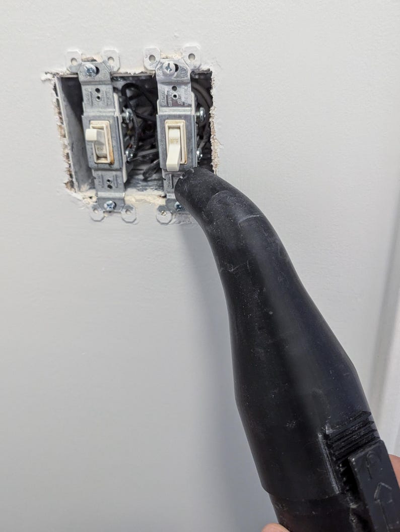 Puede incluir: Dos interruptores de luz instalados en una pared, con cableado expuesto. Una boquilla negra apunta hacia los interruptores, posiblemente para limpieza o reparaci&oacute;n. La pared est&aacute; pintada de blanco.