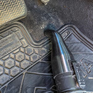 Puede incluir: Boquilla negra de aspiradora limpiando el suelo de un coche. La boquilla est&aacute; colocada cerca de una alfombrilla de goma negra con un patr&oacute;n geom&eacute;trico. Los pedales del coche son visibles en el fondo.