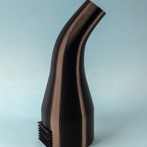 Puede incluir: Una boquilla curva negra y marr&oacute;n con una base rectangular. La boquilla tiene una superficie texturizada y un dise&ntilde;o curvo. La base tiene una serie de l&iacute;neas paralelas. El objeto est&aacute; sobre un fondo azul claro.
