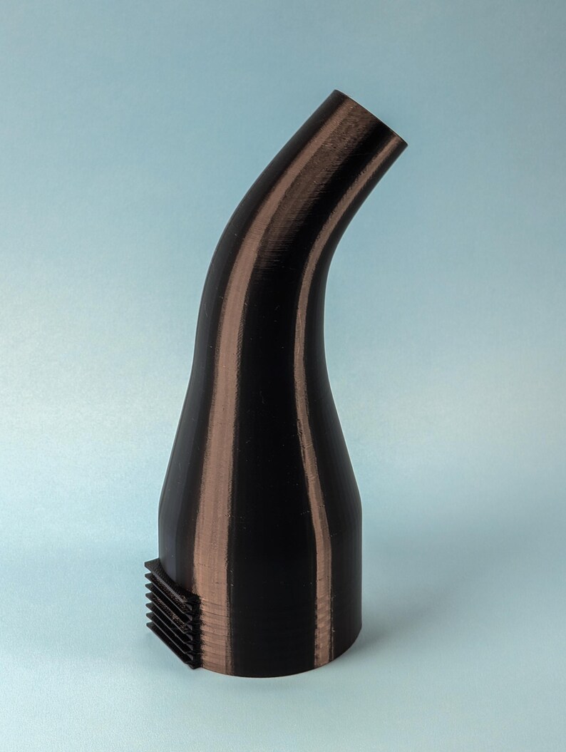Puede incluir: Una boquilla curva negra y marr&oacute;n con una base rectangular. La boquilla tiene una superficie texturizada y un dise&ntilde;o curvo. La base tiene una serie de l&iacute;neas paralelas. El objeto est&aacute; sobre un fondo azul claro.