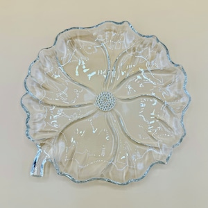 May include: Clear glass serving dish in a flower shape. The dish has a silver rim and a silver gem-like centre. The glass has etched floral designs. The dish is on a light beige background.