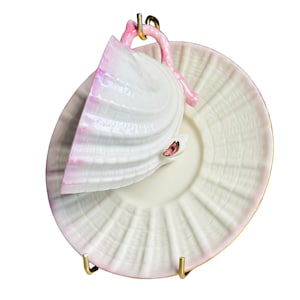 May include: A white teacup and saucer set with a seashell design. The teacup is shaped like a seashell with a pink handle and rim. The saucer is white with a ridged pattern and a gold rim. The set is displayed on a gold stand.