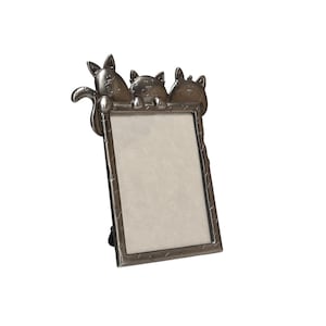 May include: A silver-coloured metal picture frame featuring three cat figures. The cats are detailed and sit above a rectangular frame with a light-coloured interior. The frame has a decorative edge and is designed to hold a photograph.