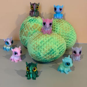 Könnte beinhalten: Eine Sammlung farbenfroher, miniaturhafter Drachenfiguren mit großen, glitzernden Augen. Die Drachen sind in verschiedenen Farben erhältlich, darunter Rosa, Blau, Grün und Lila. Sie werden auf grünen, strukturierten Kugeln mit goldenen Glitzerakzenten präsentiert.