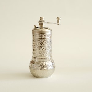 May include: A silver pepper mill with an engraved floral pattern. The mill features a cylindrical body, a rounded base, and a crank handle. The design is consistent, creating a decorative kitchen tool. The mill is made of metal.