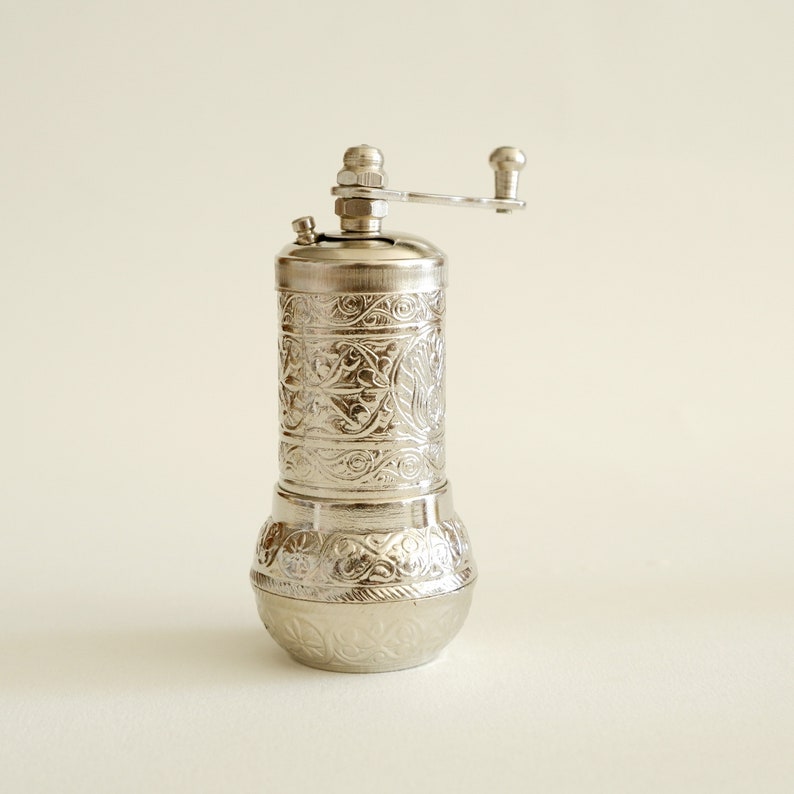 May include: A silver pepper mill with an engraved floral pattern. The mill features a cylindrical body, a rounded base, and a crank handle. The design is consistent, creating a decorative kitchen tool. The mill is made of metal.