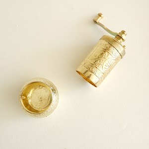 May include: A gold-coloured, vintage-style pepper grinder with an ornate floral design. The grinder is open, revealing the interior. The lid is also gold and has a matching floral pattern. The items are on a cream-coloured surface.