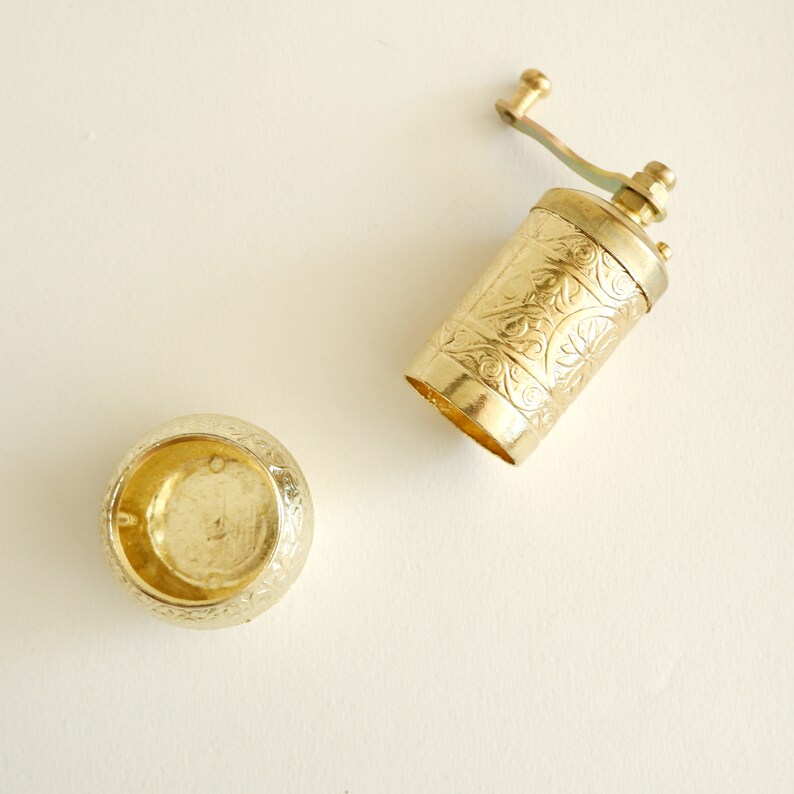 May include: A gold-coloured, vintage-style pepper grinder with an ornate floral design. The grinder is open, revealing the interior. The lid is also gold and has a matching floral pattern. The items are on a cream-coloured surface.
