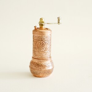 May include: Copper pepper mill with detailed floral and scrollwork. The mill has a brass top and a silver-coloured crank handle. A decorative kitchen accessory.