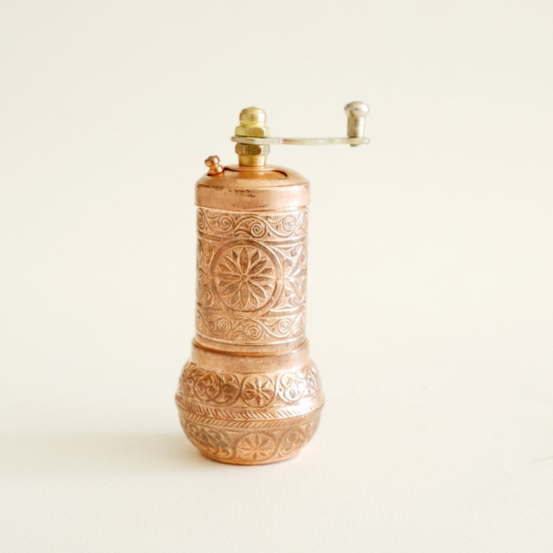 May include: Copper pepper mill with detailed floral and scrollwork. The mill has a brass top and a silver-coloured crank handle. A decorative kitchen accessory.