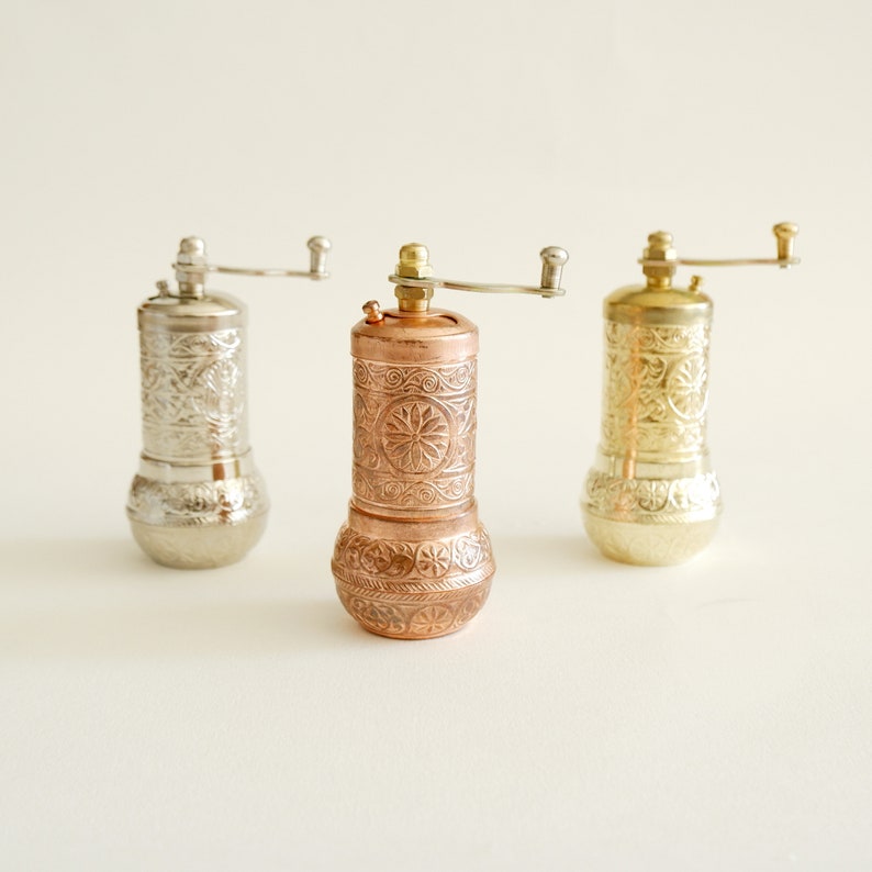 May include: Three decorative pepper grinders in silver, copper, and gold. Each grinder features an ornate, embossed floral pattern and a hand crank. The grinders are cylindrical with a rounded base.