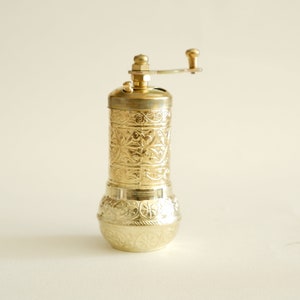 May include: A gold-coloured pepper mill with an ornate, engraved floral pattern. The mill has a cylindrical body, a rounded base, and a crank handle on top. The design is consistent throughout the mill, creating a cohesive and decorative kitchen tool.