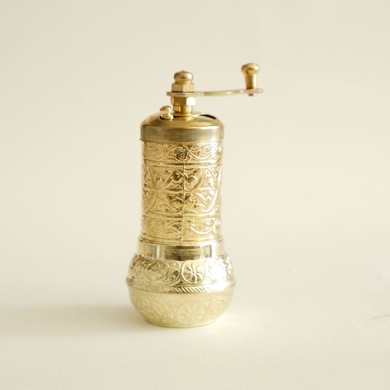 May include: A gold-coloured pepper mill with an ornate, engraved floral pattern. The mill has a cylindrical body, a rounded base, and a crank handle on top. The design is consistent throughout the mill, creating a cohesive and decorative kitchen tool.