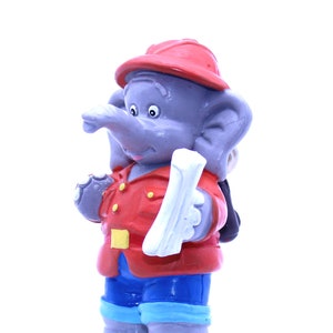 Könnte beinhalten: Eine graue Elefantenfigur, die einen roten Hut und eine rote Jacke mit einem gelben Knopf trägt. Der Elefant hält ein weißes Papier in seiner rechten Hand und trägt eine blaue Hose.