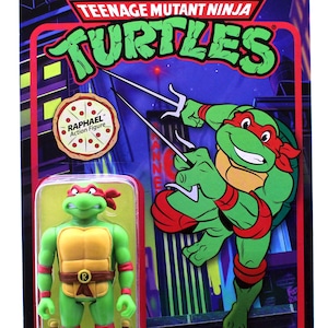 Könnte beinhalten: Teenage Mutant Ninja Turtles Actionfigur von Raphael. Die Figur ist grün mit einem roten Bandana, einer gelben Schale und einem braunen Gürtel. Er hält zwei Sai-Waffen. Die Figur ist in einer Retro-Kartonverpackung mit einem Cartoon-Bild von Raphael auf der Rückseite verpackt.