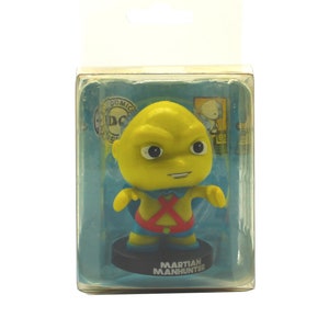 Puede incluir: Una figura de acción amarilla de Martian Manhunter en un paquete de plástico. La figura lleva un traje rojo y azul con una X en el pecho. El paquete tiene un fondo azul con el logotipo de DC Comics y las palabras "Martian Manhunter".