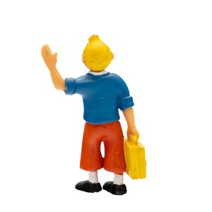 May include: A figurine of a young person with yellow hair, wearing a blue shirt, orange shorts, and black shoes. The figure is waving with one hand and holding a yellow suitcase in the other.