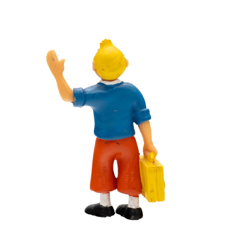 May include: A figurine of a young person with yellow hair, wearing a blue shirt, orange shorts, and black shoes. The figure is waving with one hand and holding a yellow suitcase in the other.