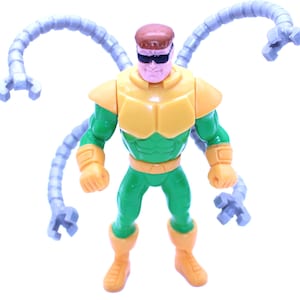 May include: A plastic action figure of Doctor Octopus, a comic book villain. The figure is primarily green and yellow, with four articulated, gray mechanical arms extending from its back. The figure wears black sunglasses and has a brown head.