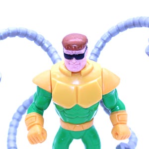 May include: A Dr. Octopus action figure. The figure is primarily green and yellow, with four articulated, light blue mechanical arms extending from its back. The character wears black sunglasses and has a brown hair.