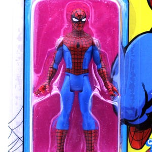 Pu&ograve; includere: Una action figure di Spider-Man rossa e blu in una confezione blister di plastica. La figura &egrave; in posa con le braccia distese e le gambe unite. La figura &egrave; su uno sfondo rosa.