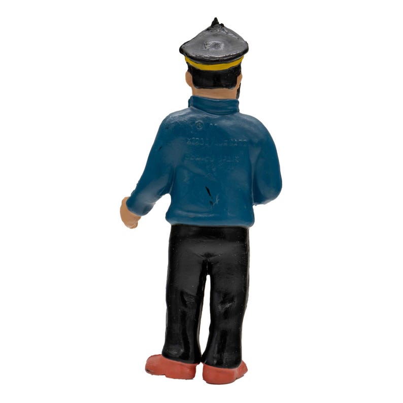 May include: A figurine of a sailor from the back. The sailor is wearing a dark blue jacket, black pants, a black hat with a yellow band, and red shoes. The figurine is standing.