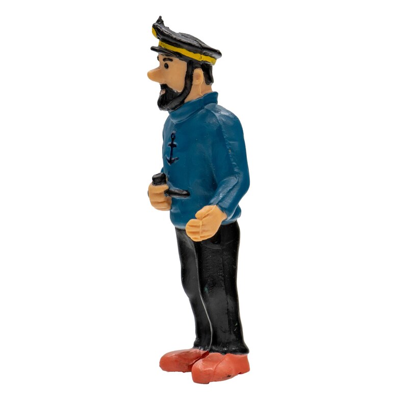 May include: A figurine of a captain in a blue sweater with an anchor emblem, black pants, and red shoes. The captain wears a black hat with a yellow band and has a black beard. He is holding a pipe.