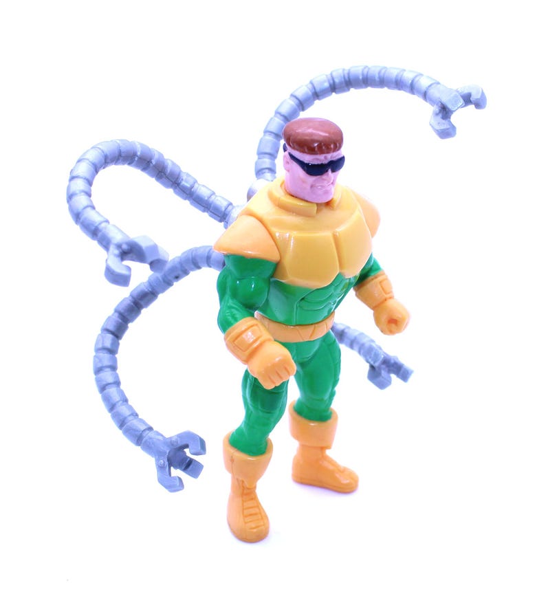 May include: A Dr. Octopus action figure. The figure is wearing a yellow and green outfit with four articulated mechanical arms. The figure has a brown head and is wearing black sunglasses. The figure is standing on a white surface.