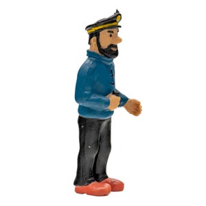 May include: A figurine of a cartoon captain, wearing a black hat with a yellow band, a blue sweater, black pants, and red shoes. The captain has a black beard and is holding a small object in his right hand. The figurine is standing on a white surface.