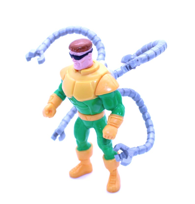 May include: A plastic action figure of a superhero with a brown head, black sunglasses, and a yellow and green suit. The figure has four articulated, gray mechanical arms extending from its back. The figure is standing on a white surface.