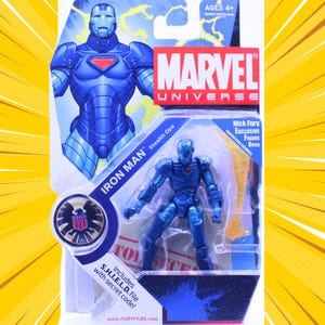 K&ouml;nnte beinhalten: Blaue Iron Man Actionfigur aus dem Marvel-Universum, verpackt auf einer gelb-blauen Karte. Die Figur befindet sich in einer dynamischen Pose, mit den Worten "IRON MAN Stealth Ops" und "Nick Fury Exclusive Figure Offer" auf der Verpackung. Enth&auml;lt eine S.H.I.E.L.D.-Datei mit Geheimcode.