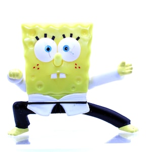 Könnte beinhalten: Gelbe Comicfigur in einem weißen Karate-Anzug und schwarzen Hosen in einer Karate-Pose. Die Figur hat blaue Augen und ein breites Lächeln.