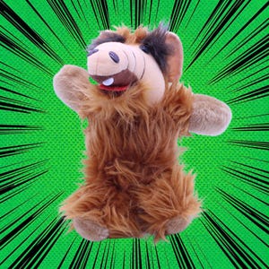 Op de afbeelding: Een pluche handpop van het personage Alf, met een beige gezicht, bruine vacht en een rode kraag. De pop heeft grote oren en staat tegen een groene achtergrond met zwarte radiale lijnen.