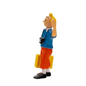 May include: A figurine of a young man with blond hair, wearing a blue shirt, orange pants, and black shoes. He carries a yellow suitcase and a camera. The figure is in a side profile, with one hand raised.