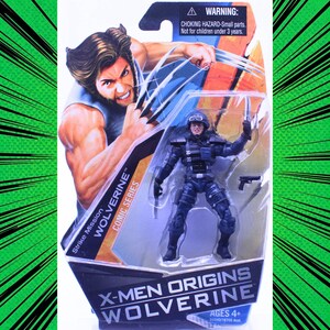 Könnte beinhalten: Eine Wolverine-Actionfigur aus der X-Men Origins Comic-Serie, verpackt in einer transparenten Blisterverpackung. Die Verpackung zeigt eine Wolverine-Illustration und den Text "Strike Mission". Die Figur ist ein Soldat in schwarzer Uniform. Alter 4+.