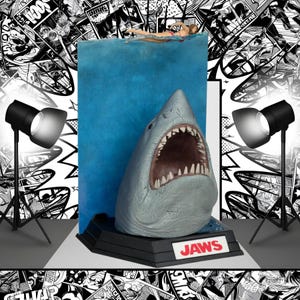 Puede incluir: Una escultura detallada de un gran tiburón blanco con la boca abierta, dientes visibles, emergiendo de agua azul. La escultura está montada sobre una base negra con la palabra "JAWS" en rojo. Una mujer nada en el agua sobre el tiburón.