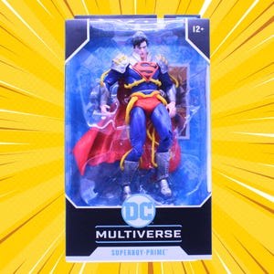 Könnte beinhalten: Eine Superboy-Prime Actionfigur in einer DC Multiverse Sichtverpackung. Die Figur trägt einen blau-roten Anzug mit rotem Umhang, silbernen Rüstungsakzenten und einem gelben Gurt. Die Verpackung zeigt das DC-Logo sowie den Text Superboy-Prime und 12+.