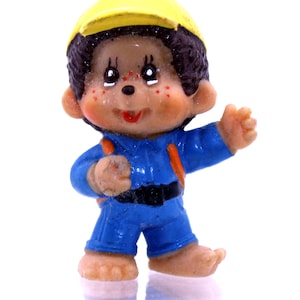 Könnte beinhalten: Eine kleine Plastikfigur eines Affen, der einen gelben Helm und eine blaue Latzhose trägt. Der Affe hat braunes Haar, eine braune Nase und ein breites Lächeln.