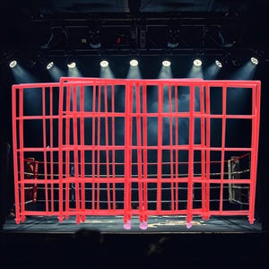 Op de afbeelding: Een felrode kooiconstructie, waarschijnlijk voor een boks- of worstelring, domineert de voorgrond. De kooi bestaat uit een raster van verticale en horizontale staven. Op de achtergrond zijn podiumverlichting en een boksring te zien.
