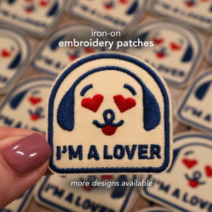 May include: Embroidered patch with a dog-like face, heart-shaped eyes, and the text "I'M A LOVER." The patch is cream-colored with blue and red accents. The words "iron-on embroidery patches" and "more designs available" are also visible.