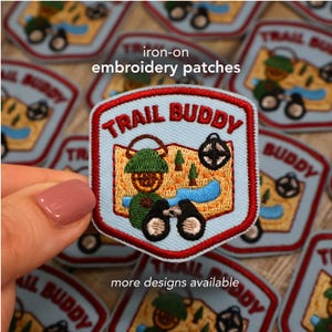 May include: Iron-on embroidery patches with a light blue background and a red border. The patches feature the text "TRAIL BUDDY" and a design of a person with binoculars, a map, and trees. The text "more designs available" is also visible.